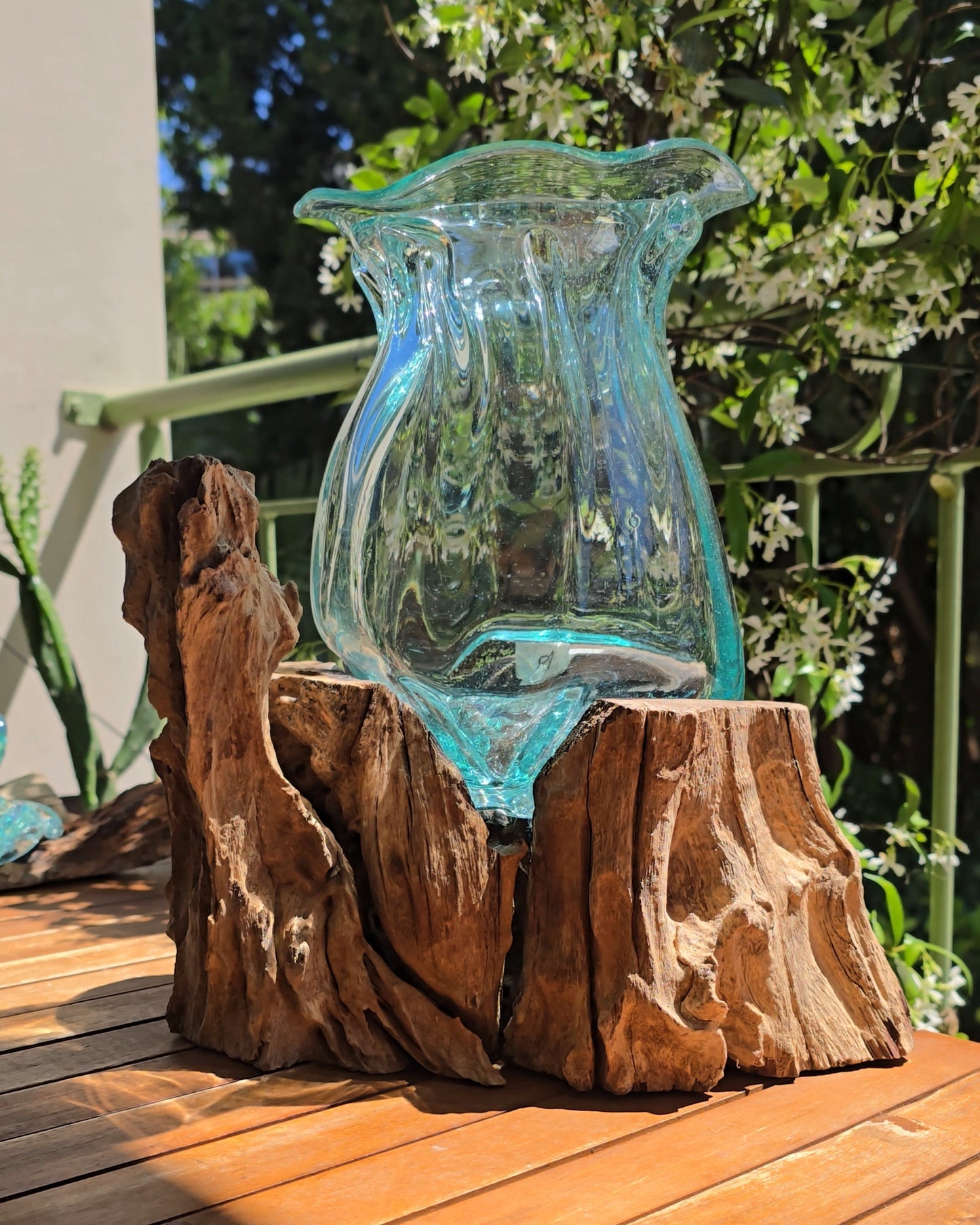 ase à lèvres artisanal 25cm vu de côté, col en verre soufflé turquoise aux bords irréguliers et organiques, socle en racine de teck aux veines apparentes, posé sur table en bois en plein air — verre de fenêtres recyclées soufflé directement sur le bois, pièce unique fait main Indonésie — LaPailleBambou Six-Fours-les-Plages