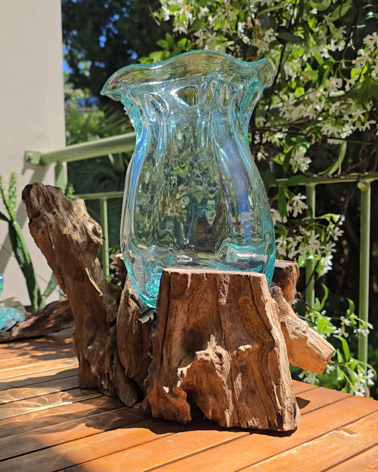 Grand vase à lèvres 25cm en verre soufflé aqua translucide posé sur racine de teck brute et nouée, ouverture ondulée formée par le souffle de l'artisan, reflets turquoise, fond végétal ensoleillé — verre recyclé soufflé à la main sur bois, pièce unique artisanale Indonésie — LaPailleBambou Six-Fours-les-Plages
