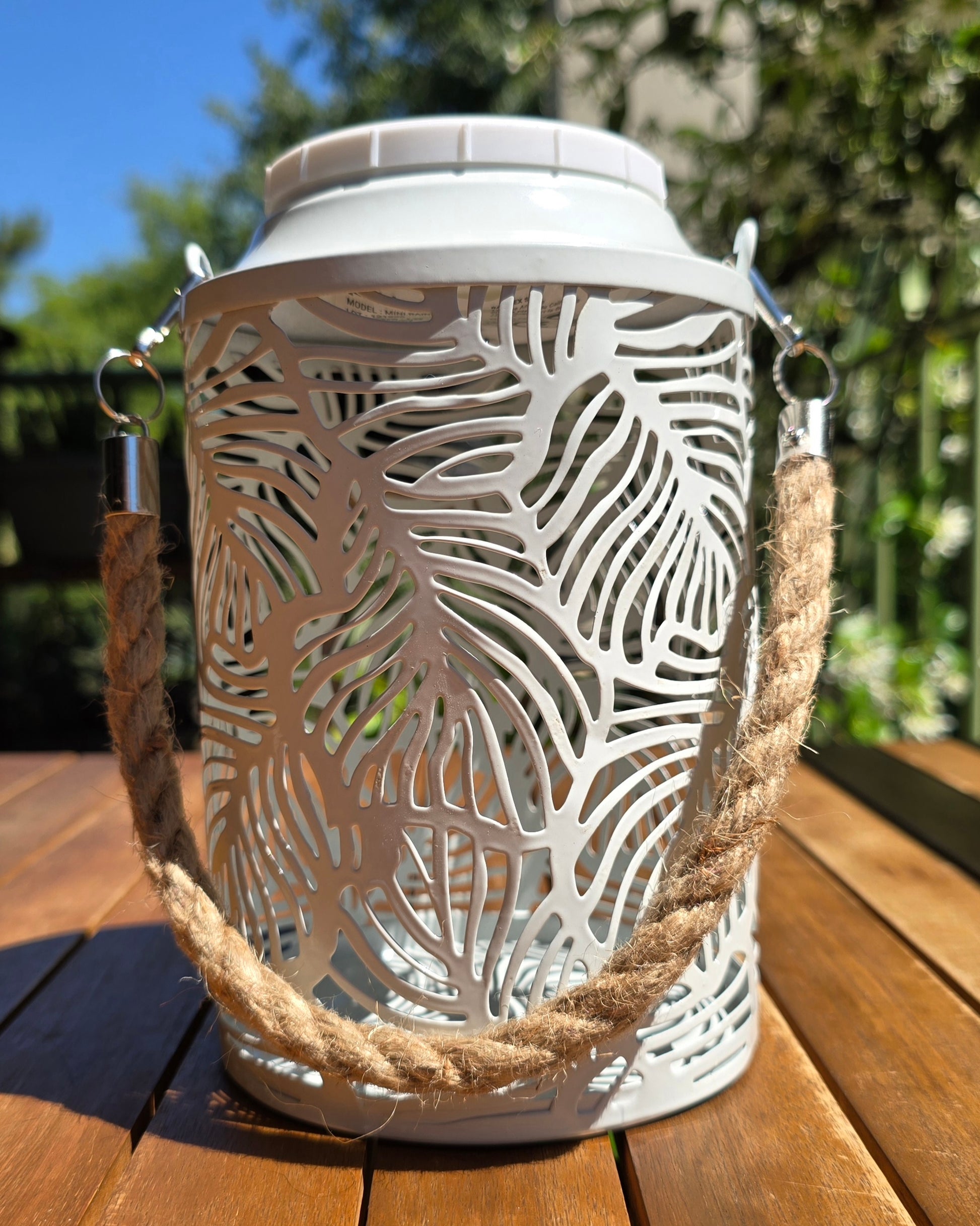Lanterne solaire Mini Rainbow Lumisky vue de trois quarts, métal blanc perforé motifs feuilles tropicales et poignée en corde naturelle, fond de jardin ensoleillé