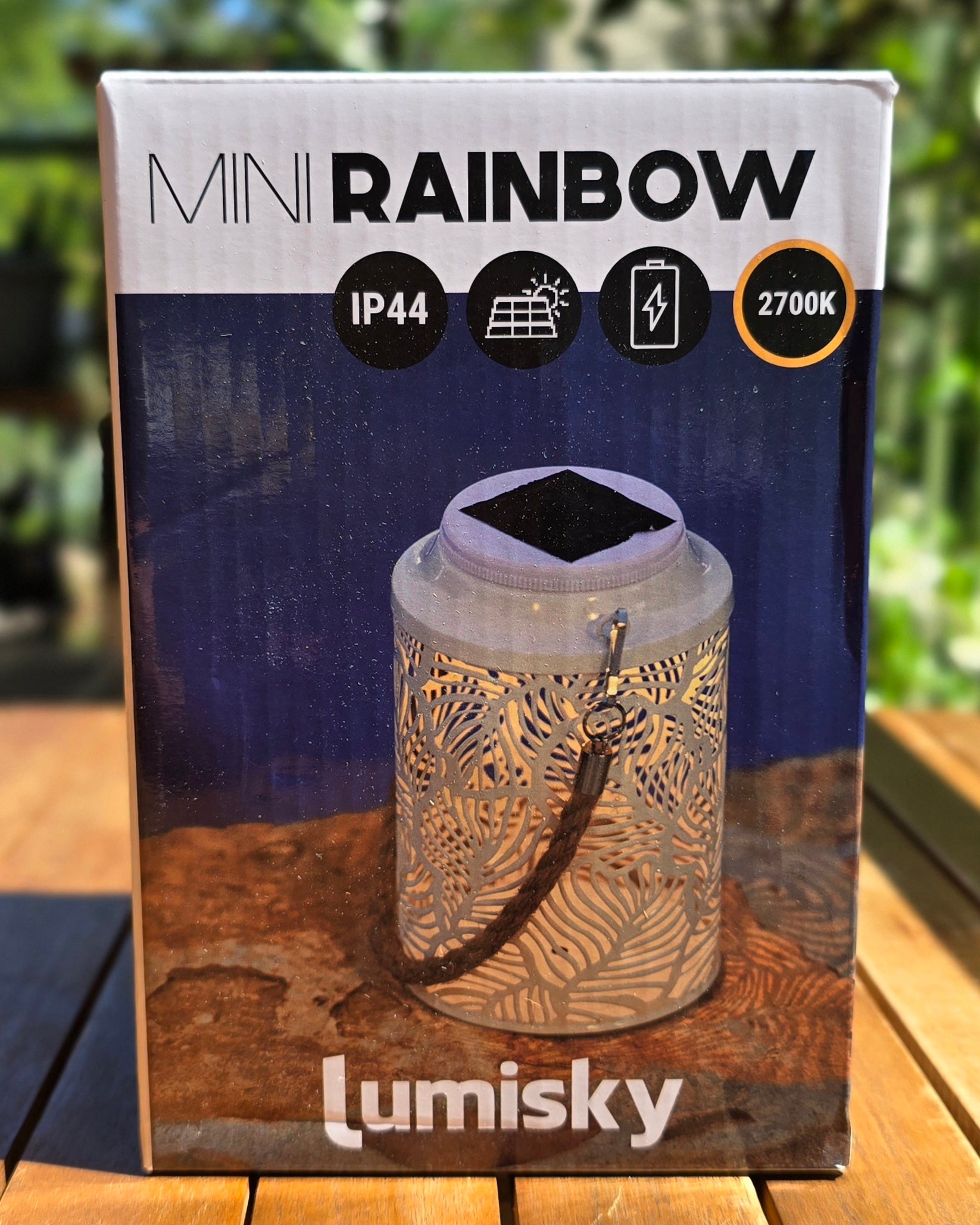Lanterne solaire Mini Rainbow Lumisky vue de face, col en métal blanc ajouré aux motifs de feuilles, poignée corde suspendue, fond de verdure extérieure