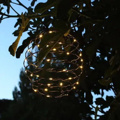 Suspension solaire ORION Lumisky suspendue à une branche d'arbre au crépuscule, micro-LEDs allumées en blanc chaud, structure tourbillon métallique, atmosphère féérique