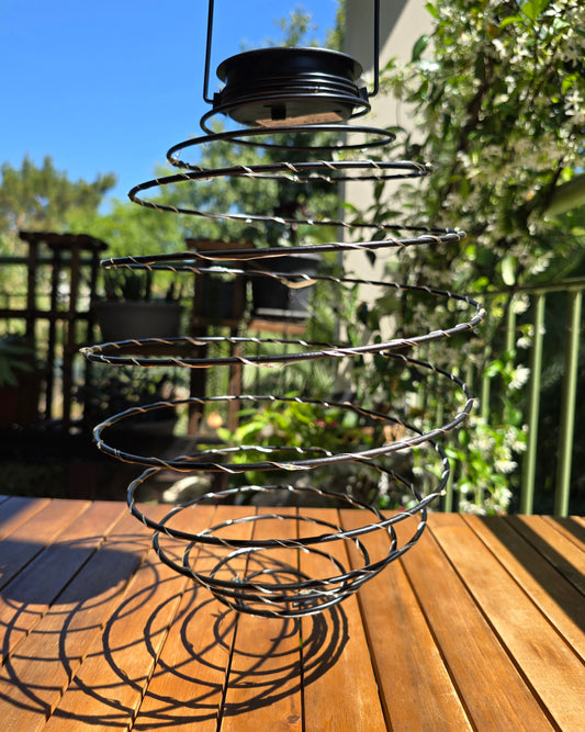 Suspension solaire ORION Lumisky vue de jour en extérieur, structure tourbillon en fil métallique tressé suspendue à un crochet, fond de terrasse et jardin verdoyant
