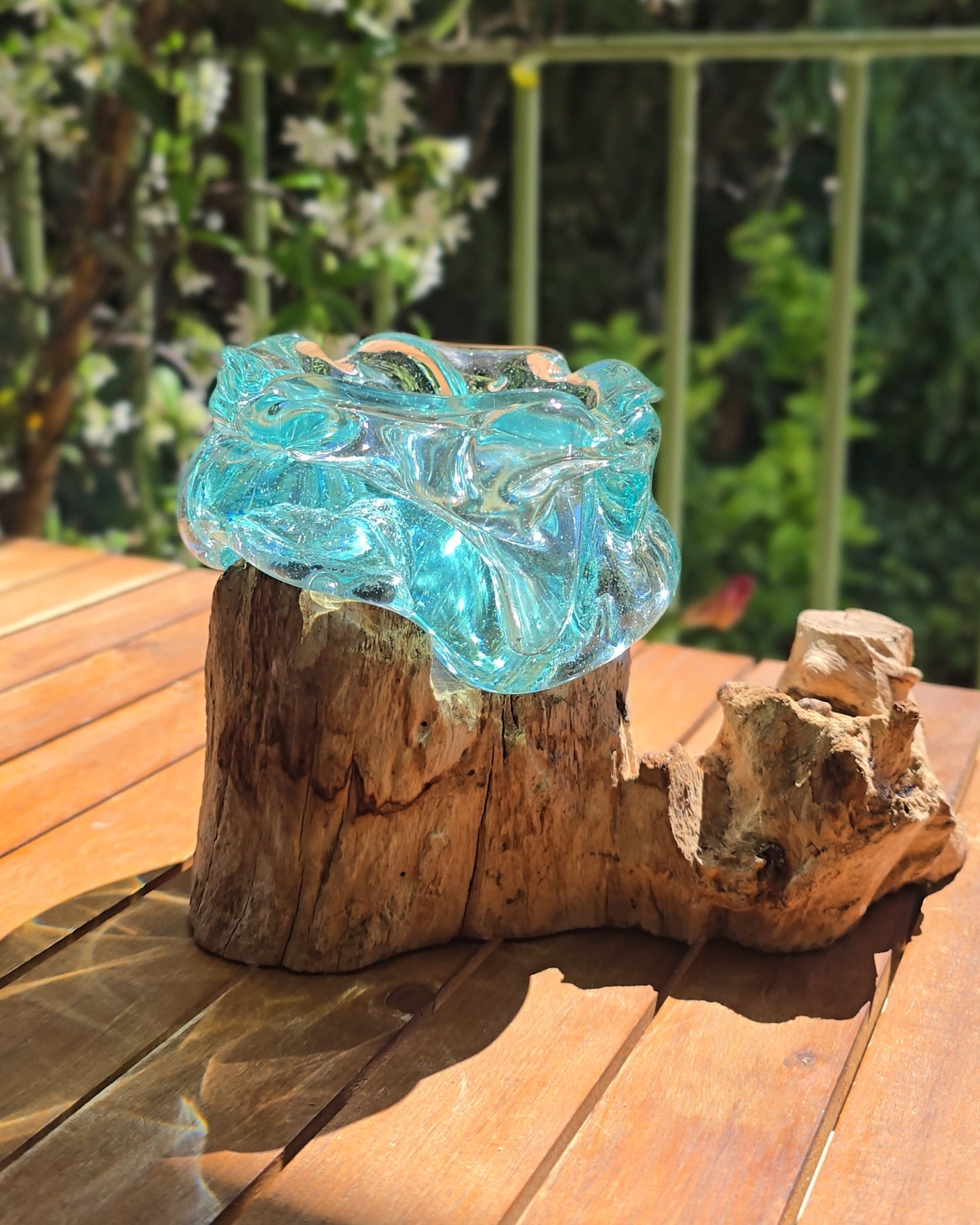 Cendrier artisanal 16cm en verre soufflé aqua posé sur souche de teck brute et nouée, ouverture arrondie aux bords organiques formés par le souffle de l'artisan, reflets turquoise translucides, fond de feuillage ensoleillé — verre de fenêtres recyclées soufflé à la main directement sur racine de bois, pièce unique Indonésie — LaPailleBambou Six-Fours-les-Plages