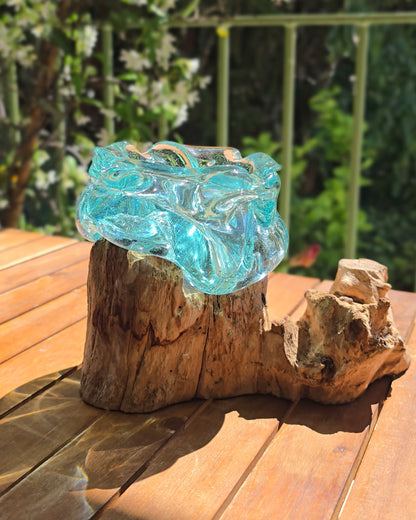 Cendrier artisanal 16cm en verre soufflé aqua posé sur souche de teck brute et nouée, ouverture arrondie aux bords organiques formés par le souffle de l'artisan, reflets turquoise translucides, fond de feuillage ensoleillé — verre de fenêtres recyclées soufflé à la main directement sur racine de bois, pièce unique Indonésie — LaPailleBambou Six-Fours-les-Plages