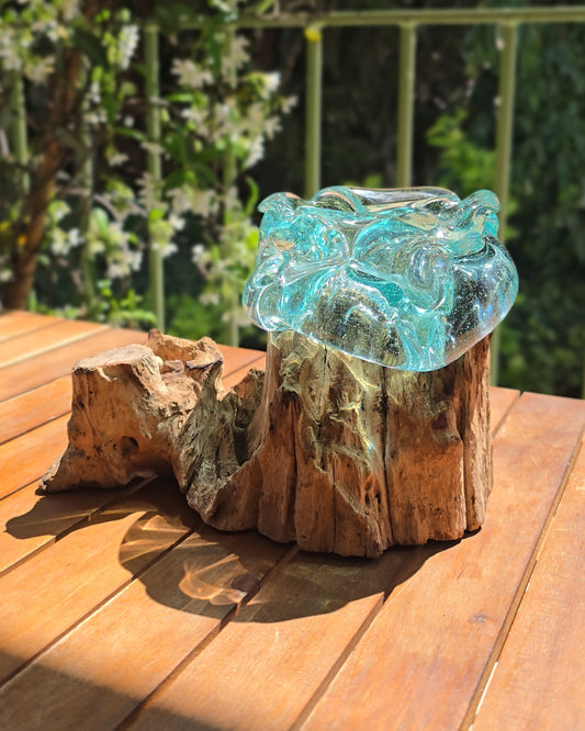 Cendrier bois et verre soufflé turquoise 12×10×16cm vu de côté, coupe en verre aqua aux contours irréguliers et vivants posée sur tronçon de racine de teck aux veines apparentes, posé sur table en bois en plein air — verre recyclé soufflé à la main sur bois gamal, artisanat fait main Indonésie — LaPailleBambou Six-Fours-les-Plages