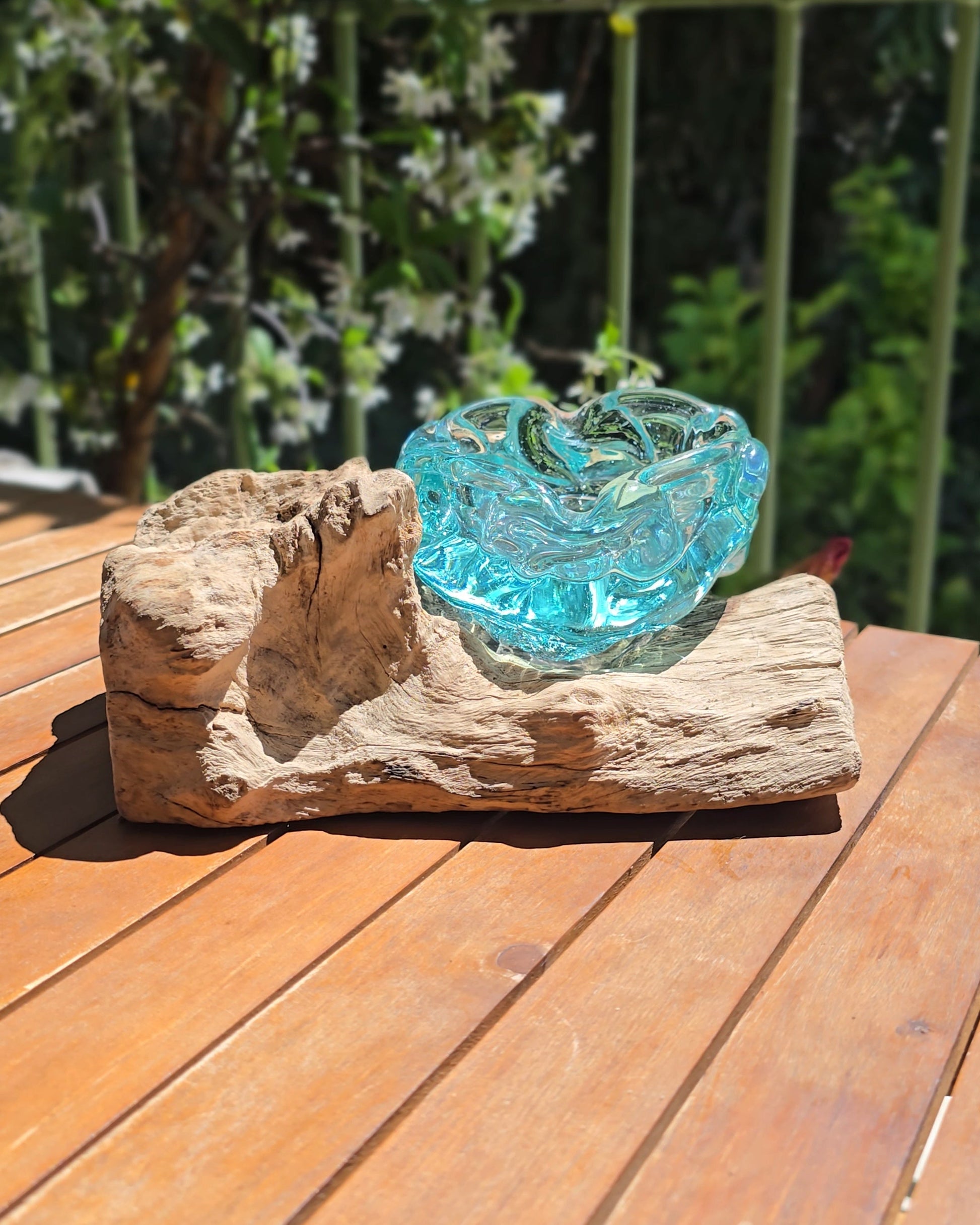 Grand cendrier artisanal 20cm en verre soufflé aqua posé sur tronc de teck brut et massif, coupe en verre turquoise translucide aux bords ondulés et organiques formés par le souffle de l'artisan, posé en plein air sur table en bois — verre de fenêtres recyclées soufflé à la main sur racine de bois gamal, pièce unique Indonésie — LaPailleBambou Six-Fours-les-Plages