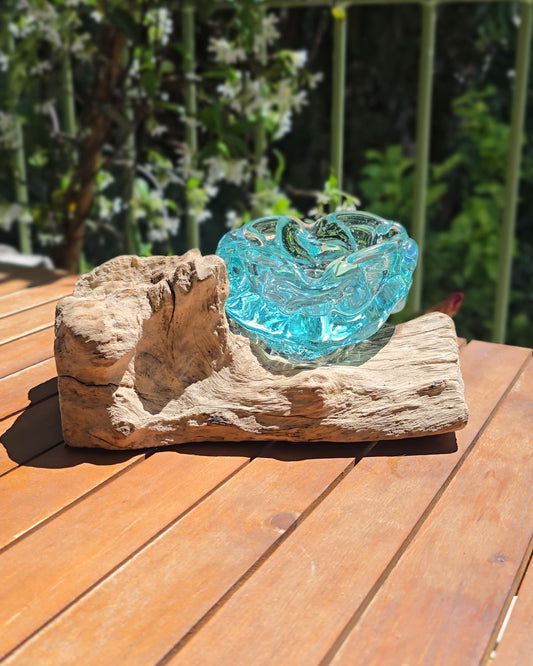 Grand cendrier artisanal 20cm en verre soufflé aqua posé sur tronc de teck brut et massif, coupe en verre turquoise translucide aux bords ondulés et organiques formés par le souffle de l'artisan, posé en plein air sur table en bois — verre de fenêtres recyclées soufflé à la main sur racine de bois gamal, pièce unique Indonésie — LaPailleBambou Six-Fours-les-Plages