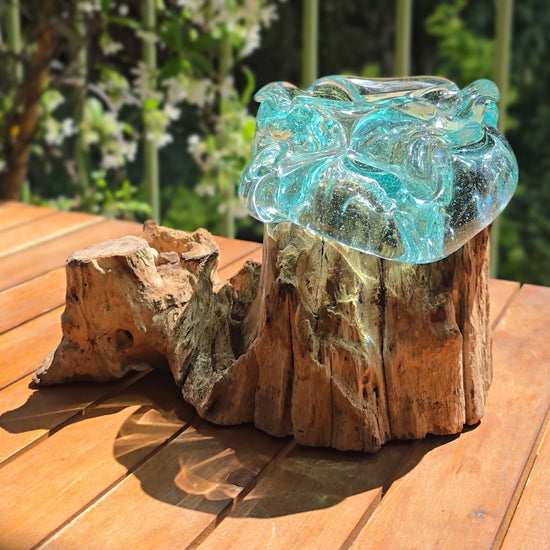 Vidéo — Gros plan du petit cendrier artisanal LaPailleBambou en verre soufflé recyclé sur socle en teck, présenté sur une table en bois au soleil, reflets aqua translucides — LLX ConceptStore