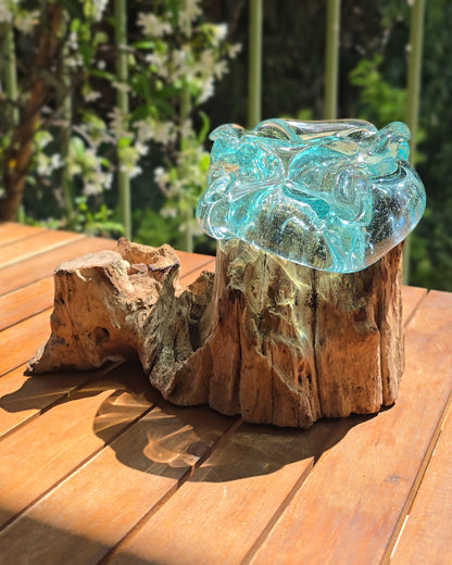 Vidéo — Gros plan du petit cendrier artisanal LaPailleBambou en verre soufflé recyclé sur socle en teck, présenté sur une table en bois au soleil, reflets aqua translucides — LLX ConceptStore