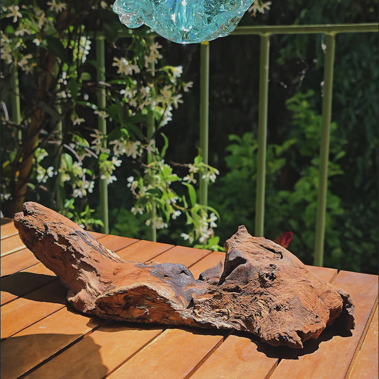 Vidéo — Gros plan du soliflore artisanal LaPailleBambou en verre soufflé recyclé sur socle en teck, posé sur une table en bois au soleil, reflets aqua translucides — LLX ConceptStore