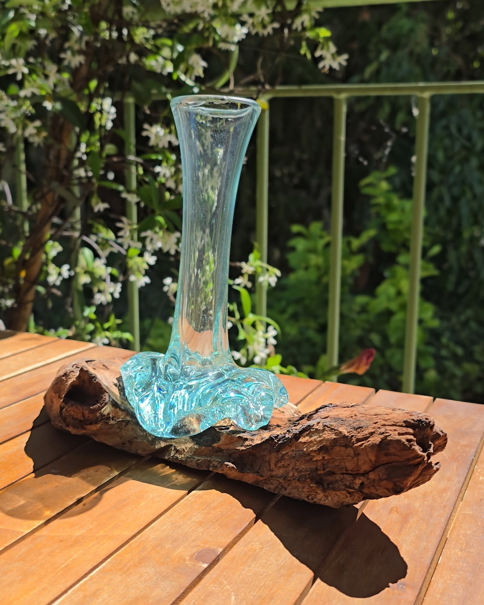 Soliflore artisanal 12cm en verre soufflé aqua élancé, col fin et transparent s'élevant depuis une racine de teck brute et nouée, reflets turquoise translucides captant la lumière du soleil, posé sur table en bois en plein air — verre recyclé soufflé à la main directement sur bois teck, pièce unique Indonésie — LaPailleBambou Six-Fours-les-Plages