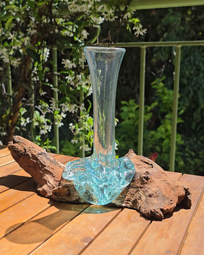 Soliflore bois et verre soufflé turquoise 19×12×21cm vu de côté, col en verre aqua élancé aux parois fines et légères posé sur souche de teck aux veines profondes et textures organiques, fond de feuillage fleuri — verre de fenêtres recyclées soufflé à la main sur racine de bois gamal, artisanat fait main Indonésie — LaPailleBambou Six-Fours-les-Plages